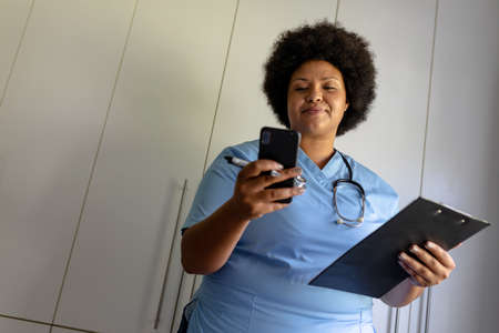 African american mid adult female nurse using smart phone while holding clipboard in hospital....