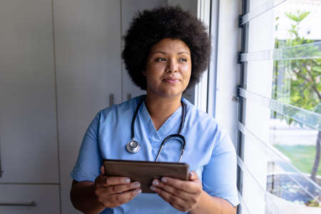 Smiling african american mid adult female nurse looking away while holding digital tablet....