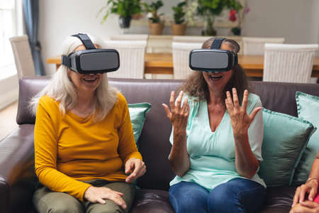 Caucasian senior female friends smiling while using vr headsets in living room at home. unaltered,...