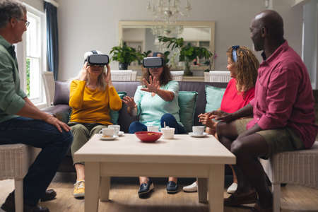 Multiracial senior woman and men with friends using vr headsets at home. unaltered, lifestyle, social gathering, friendship, virtual reality, futuristic and modern technology.の写真素材