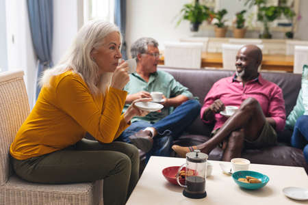 Senior woman drinking coffee while multiracial friends talking in background at home. unaltered,...