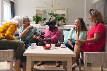 Happy multiracial senior man and women looking at male friend using vr headset at home. unaltered,...