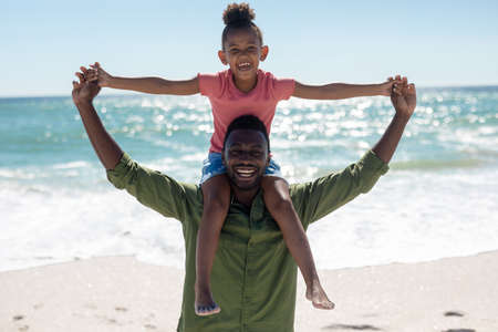 Portrait of happy african american girl sitting on father&#39;s shoulders at beach against sea. unaltered, family, lifestyle, togetherness, enjoyment and holiday concept.の写真素材