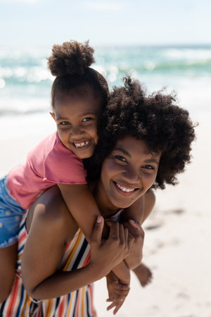 Portrait of happy african american woman giving piggyback to daughter at beach on sunny day. unaltered, family, lifestyle, togetherness, enjoyment and holiday concept.の写真素材
