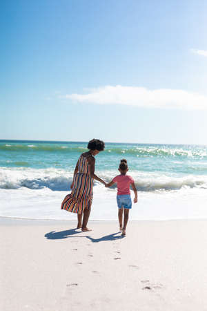 African american mother holding hand of daughter while walking towards sea at beach on sunny day....