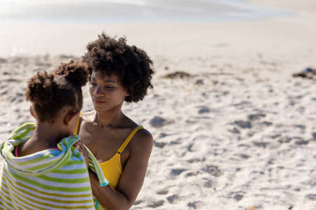 African american woman wrapping daughter with towel on sunny day at beach. unaltered, family, lifestyle, togetherness, enjoyment and holiday concept.の写真素材