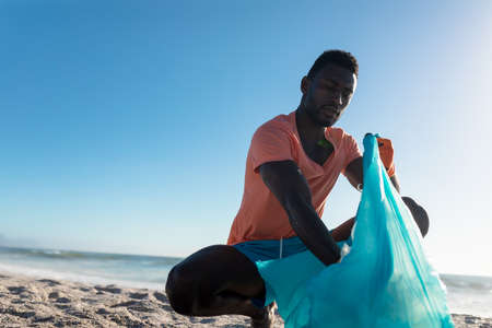 African american man crouching while collecting garbage in plastic bag at beach against blue sky. unaltered, responsibility and environmental issues concept.の写真素材