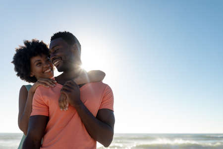 Happy back lit african american woman embracing boyfriend at beach against blue sky with copy space. unaltered, lifestyle, love, togetherness and holiday concept.の写真素材