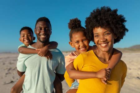 Portrait of smiling african american parents piggybacking children at beach against sky. unaltered, family, lifestyle, togetherness, enjoyment and holiday concept.の写真素材