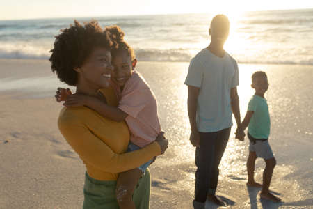 Happy african american mother carrying daughter while standing by father and son at beach. unaltered, family, lifestyle, togetherness, enjoyment and holiday concept.の写真素材