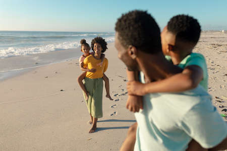 African american mother piggybacking daughter while father and son looking at them. unaltered, family, lifestyle, togetherness, enjoyment and holiday concept.の写真素材