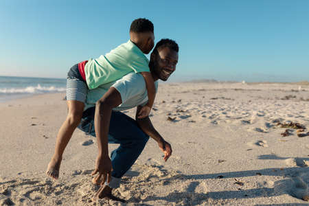 Happy african american man giving piggyback to son at beach son on sunny day. unaltered, family, lifestyle, togetherness, enjoyment and holiday concept.の写真素材