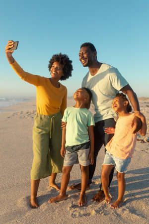 Happy african american woman taking selfie with family from smartphone at beach against blue sky. unaltered, family, lifestyle, togetherness, technology and holiday concept.の写真素材