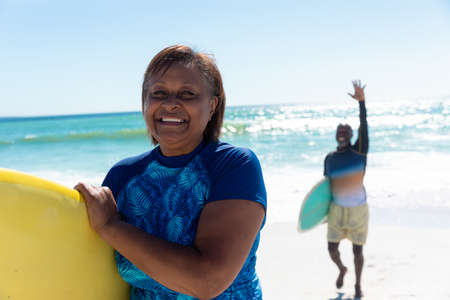 Portrait of smiling african american woman carrying surfboard with senior man waving at beach....