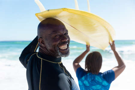 Rear view portrait of african american senior man carrying surfboard on head with woman at beach. unaltered, togetherness, active lifestyle, aquatic sport and holiday concept.の写真素材