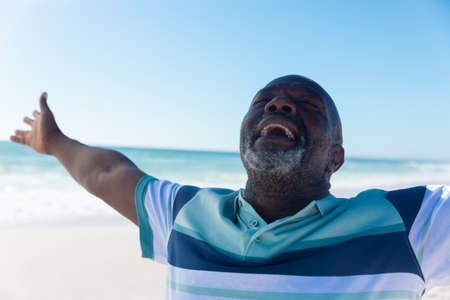 Happy african american senior retired man with eyes closed and arms outstretched at beach. unaltered, active lifestyle, enjoyment and holiday concept.の写真素材