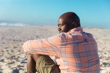 Rear view of retired african american senior man looking away while sitting at beach on sunny day. unaltered, active lifestyle, enjoyment and holiday concept.の写真素材