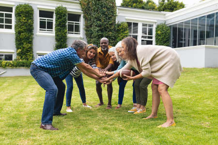 Multiracial senior male and female friends stacking hands while playing rugby in backyard....