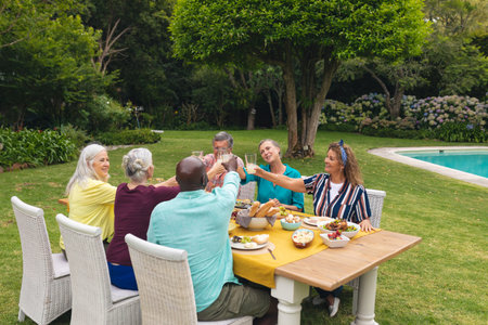 Multiracial senior male and female friends toasting wine at table during backyard party. unaltered, lifestyle, leisure, togetherness, friendship, retirement and backyard party.の写真素材