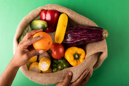 Directly above view of mixed race man hand holding fresh orange fruit over vegetables in burlap sack. unaltered, food, healthy eating, organic concept.の写真素材