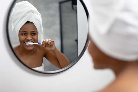 African american mid adult woman looking at mirror and brushing teeth in bathroom. unaltered, dental health, lifestyle, hygiene, routine.の写真素材