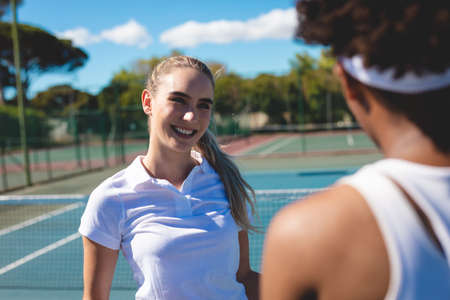 Smiling caucasian female athlete looking at african american player at tennis court on sunny day. unaltered, sport, competition and tennis game concept.の写真素材