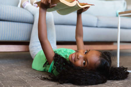 Portrait of african american elementary girl reading book while lying on floor with legs over couch....
