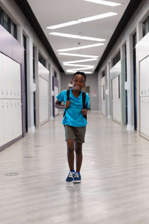 Portrait of smiling african american elementary schoolboy with backpack walking in school corridor. unaltered, education, childhood, happiness and school concept.の写真素材
