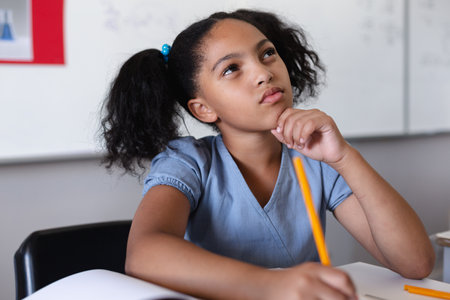 Thoughtful biracial elementary girl with hand on chin looking up while studying at desk in...