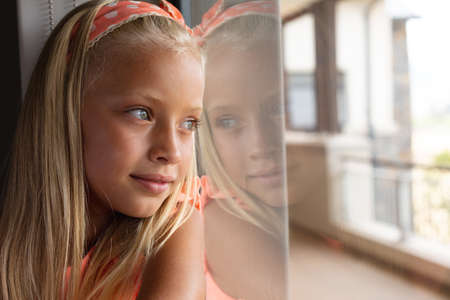 Close-up of caucasian elementary schoolgirl leaning on glass window in classroom. unaltered, education, childhood, contemplation, day dreaming, smiling and school concept.の写真素材