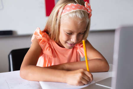 Smiling caucasian elementary schoolgirl writing on book while studying with laptop at desk in class. unaltered, education, learning, studying, concentration, wireless technology and school concept.の写真素材