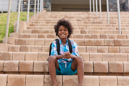 Portrait of smiling african american elementary schoolboy sitting on school building steps....