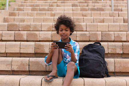 African american elementary schoolboy using smart phone while sitting on school building steps. unaltered, education, childhood, wireless technology and school concept.の写真素材