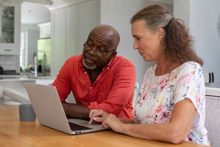 Multiracial senior couple using laptop while sitting at table in living room. unaltered, lifestyle,...