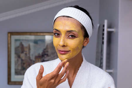 Portrait of caucasian young woman applying facial mask in bathroom. unaltered, lifestyle, skin care,...