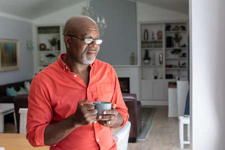 Thoughtful african american senior man having coffee while standing in living room at home. ....