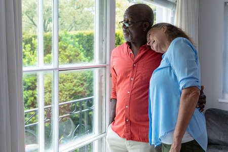 Multiracial senior couple looking through window while standing at home. unaltered, lifestyle, retirement, togetherness, contemplation.の写真素材