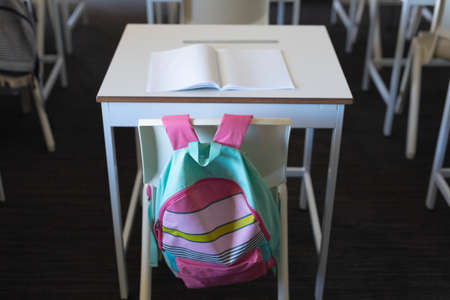Backpack hanging on chair at desk with book in classroom. unaltered, education and school concept.の写真素材