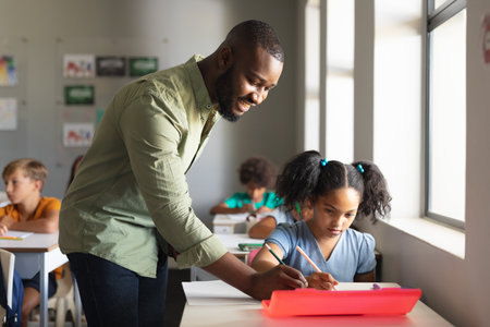 Smiling african american young male teacher assisting african american schoolgirl at desk in class. unaltered, education, learning, childhood, occupation, teaching and school concept.の写真素材