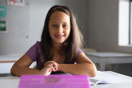 Portrait of smiling caucasian elementary schoolgirl sitting at desk in classroom. unaltered,...
