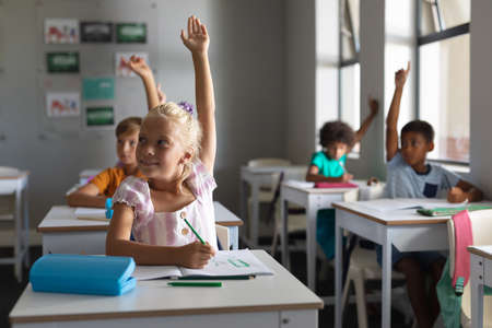 Multiracial elementary school students with hands raised sitting at desk in classroom. unaltered,...