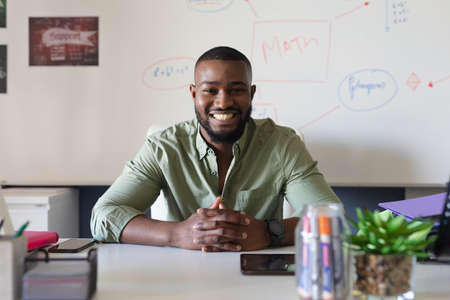 Portrait of smiling african american young male teacher sitting at desk against whiteboard in class. unaltered, education, teaching, occupation and school concept.の写真素材