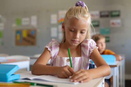 Caucasian elementary schoolgirl writing on book at desk in classroom. unaltered, teacher, education, learning, studying and school concept.の写真素材