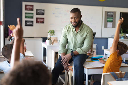 African american young male teacher looking at multiracial students raising hands during class....