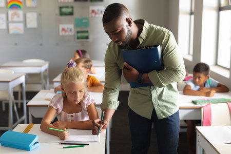 African american young male teacher assisting caucasian elementary schoolgirl at desk in classroom....