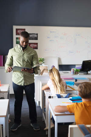African american young male teacher reading file while standing by caucasian students in classroom....