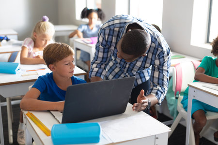 African american young male teacher teaching laptop to caucasian elementary schoolboy at desk. unaltered, education, childhood, occupation, wireless technology and school concept.の写真素材