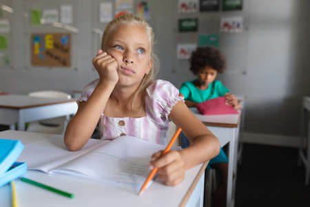 Caucasian elementary schoolgirl with hand on chin looking away while studying at desk in classroom. unaltered, education, learning, childhood, studying and school concept.の写真素材