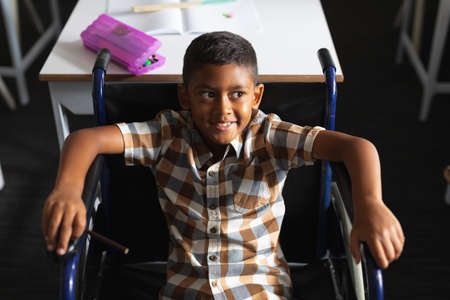 High angle view of smiling biracial elementary boy sitting on wheelchair in classroom. unaltered, education, childhood, disability, learning, physical disability and school concept.の写真素材