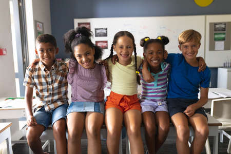 Portrait of smiling multiracial elementary school students sitting with arm around in classroom....